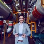 Handsome smiling supervisor in gray suit holding tablet while looking at camera. Energy plant interior.